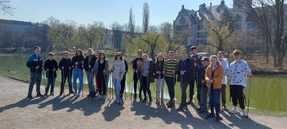 Grupa uczniów stojąca z kijkami do Nordic walking. W tle widać staw oraz budynki.